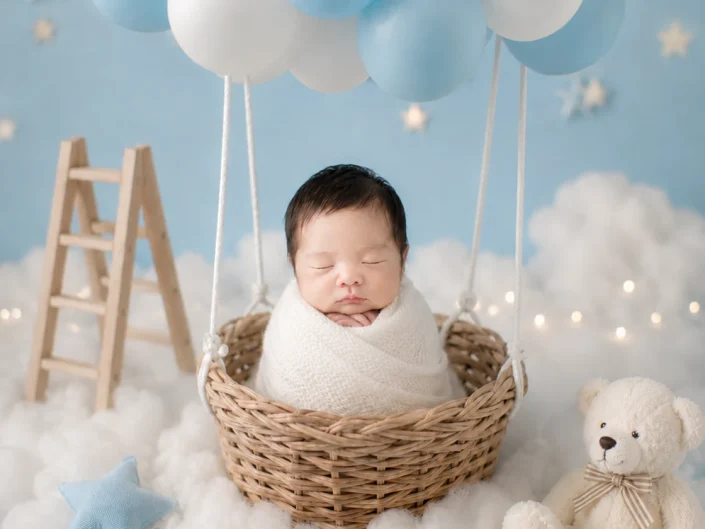 Sleeping newborn wrapped in white in a balloon basket beneath blue and white balloons at Sweetlife Photography North Lakes