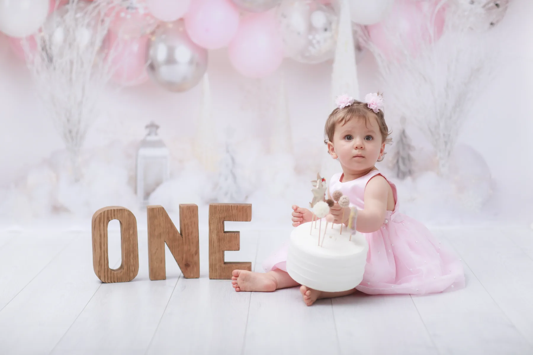Baby in a pink dress seated beside a first-birthday cake and wooden ONE letters