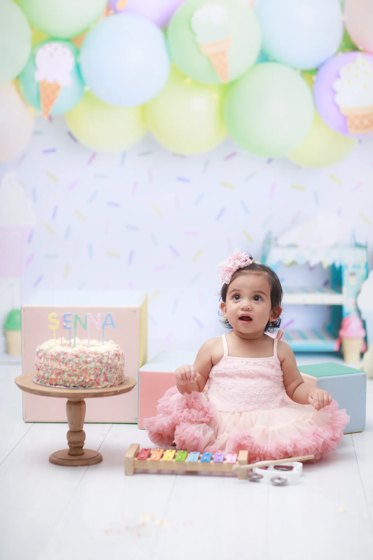 Baby in a pink dress seated beside a colourful first-birthday cake and musical toys